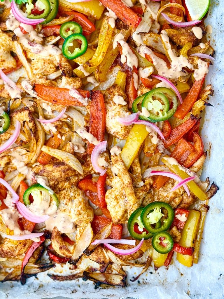 Overhead shot of roasted chicken fajitas on a sheet pan with red and yellow peppers, onions, and chipotle crema, topped with tangy pickled jalapeños and red onions.