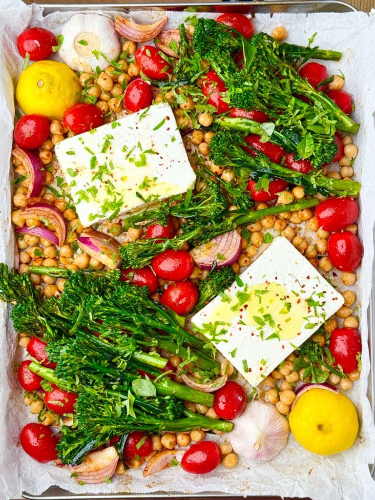 Raw ingredients for sheet pan baked feta arranged on parchment—broccolini, cherry tomatoes, red onion, chickpeas, feta, and lemon halves