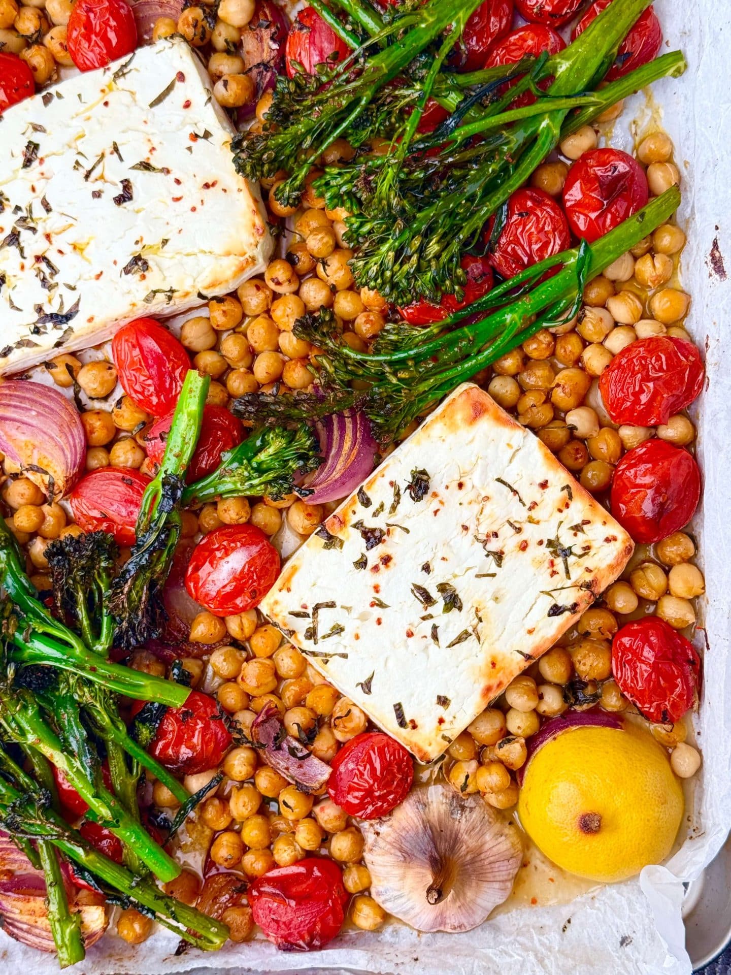 Sheet pan filled with roasted chickpeas, blistered tomatoes, broccolini, red onions, garlic, lemon, and golden baked feta