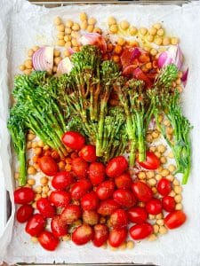 Seasoned chickpeas, tomatoes, broccolini, and red onion spread on a sheet pan before baking