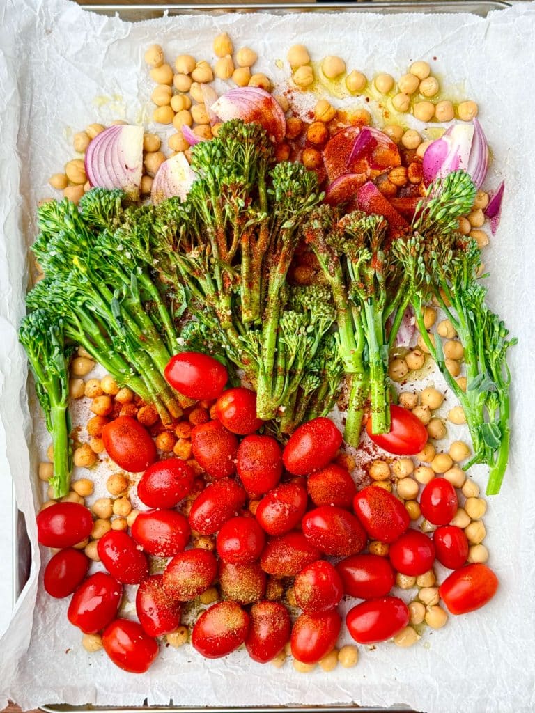 Seasoned chickpeas, tomatoes, broccolini, and red onion spread on a sheet pan before baking