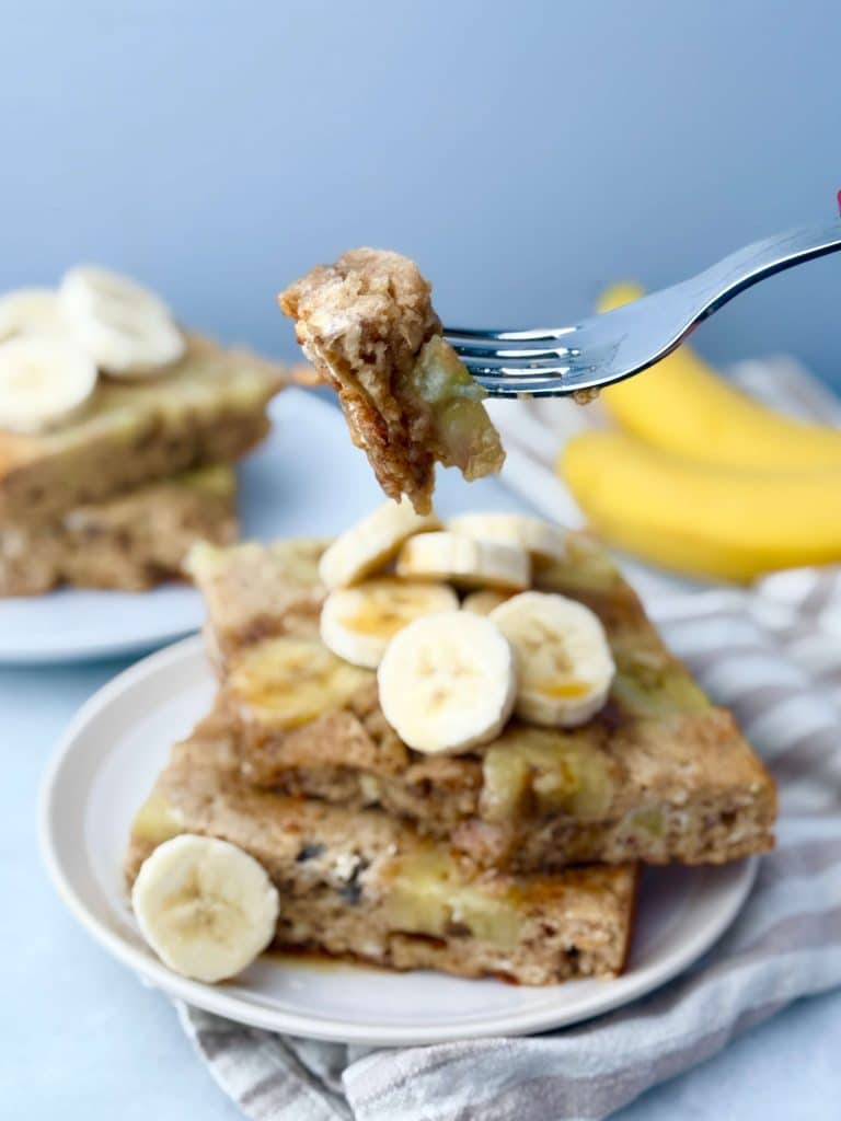 Bite of banana protein pancake on a fork with banana slices and syrup in the background.