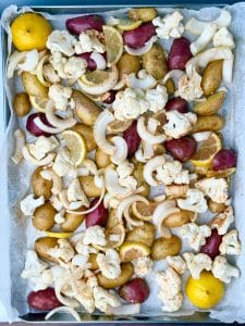 Overhead view of seasoned potatoes, cauliflower, onion, and lemon slices arranged on a parchment-lined baking sheet.