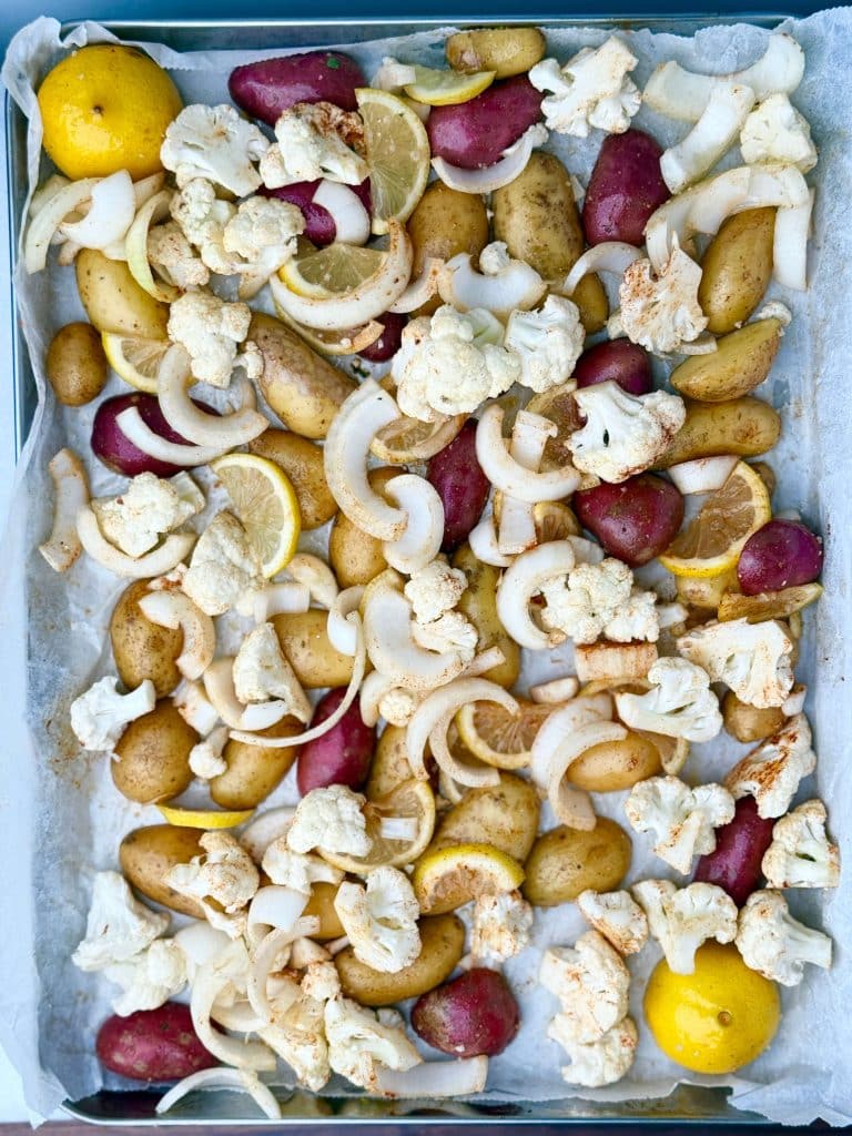 Overhead view of seasoned potatoes, cauliflower, onion, and lemon slices arranged on a parchment-lined baking sheet.