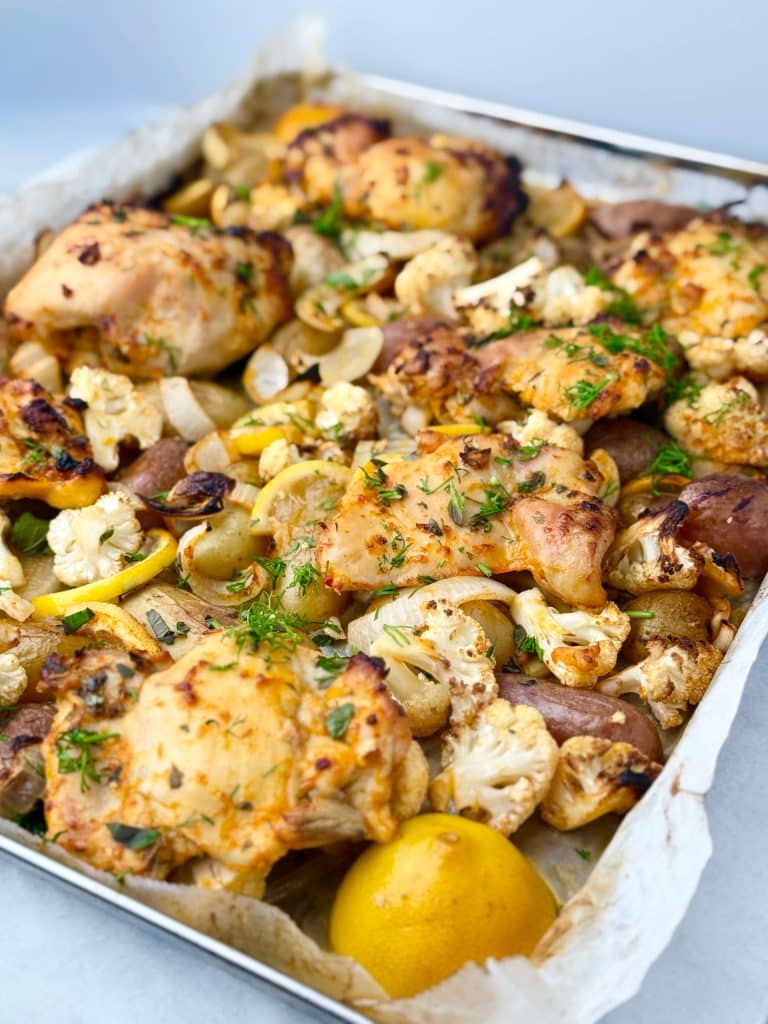 Angled sheet pan shot of roasted chicken thighs, potatoes, and cauliflower with fresh herbs and lemon halves.