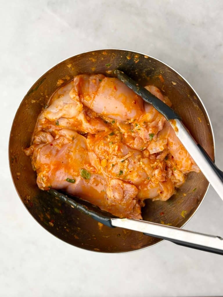 Close-up of chicken thighs marinating in lemon, garlic, and herb mixture in a stainless steel bowl.
