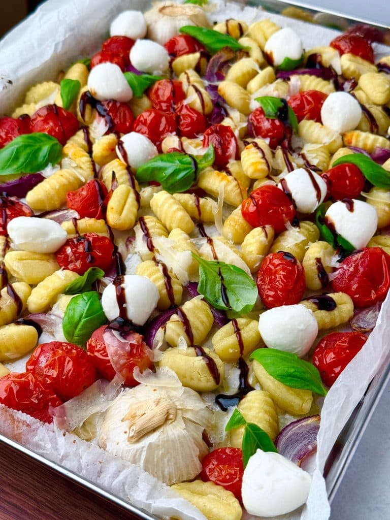 Close-up of roasted caprese gnocchi on a sheet pan with blistered cherry tomatoes, mozzarella pearls, basil leaves, and halved garlic.