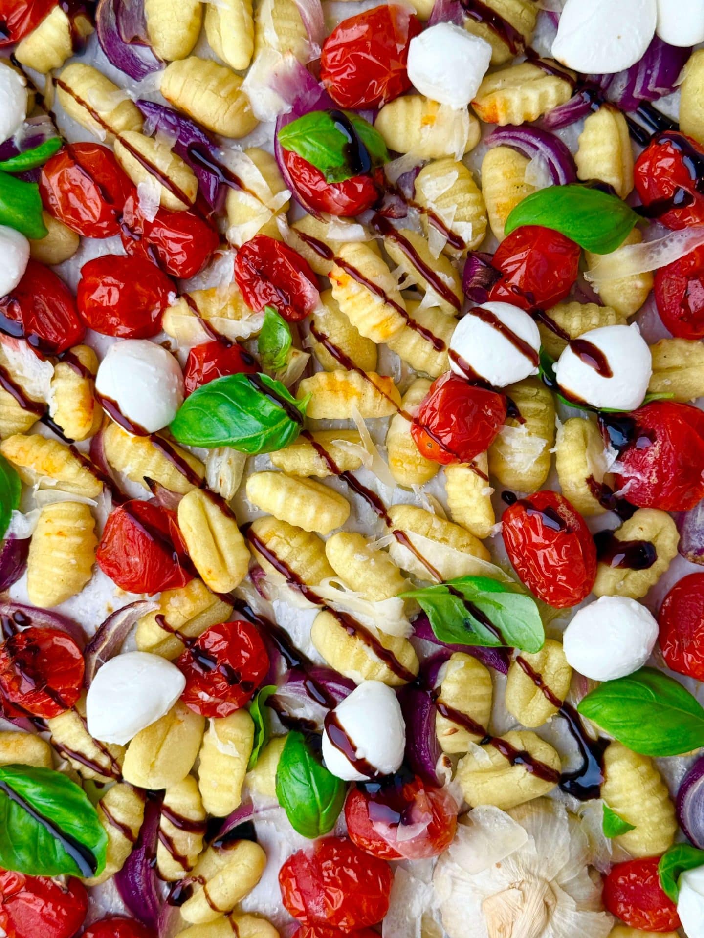 Overhead view of roasted sheet pan caprese gnocchi with cherry tomatoes, mozzarella pearls, fresh basil, and balsamic drizzle.