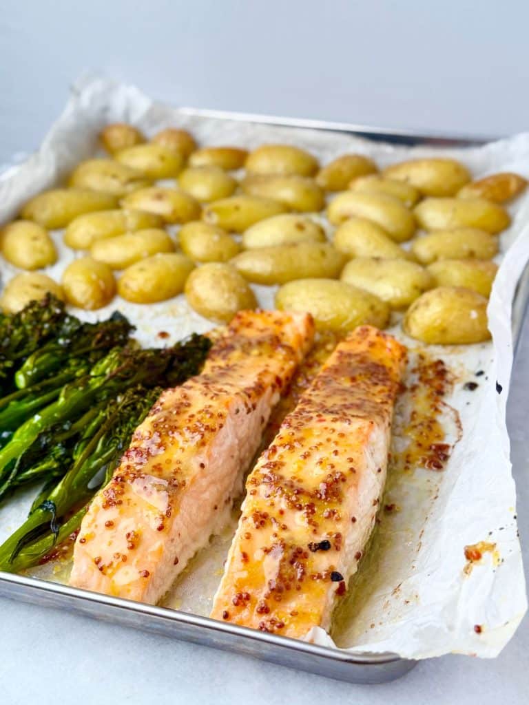 Angled shot of a sheet pan featuring two cooked honey mustard salmon fillets, roasted broccolini, and crispy baby potatoes.
