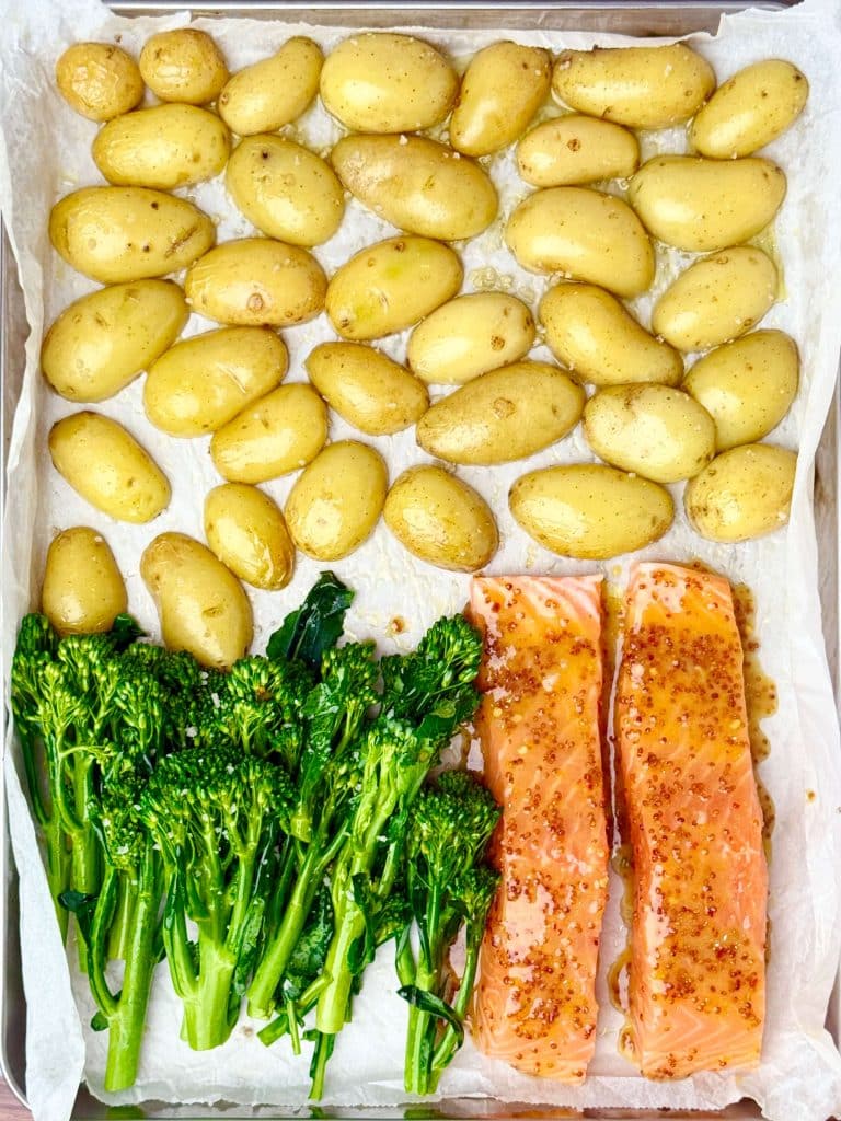Partially roasted sheet pan with golden potatoes, fresh broccolini, and raw honey mustard-glazed salmon fillets arranged side by side.