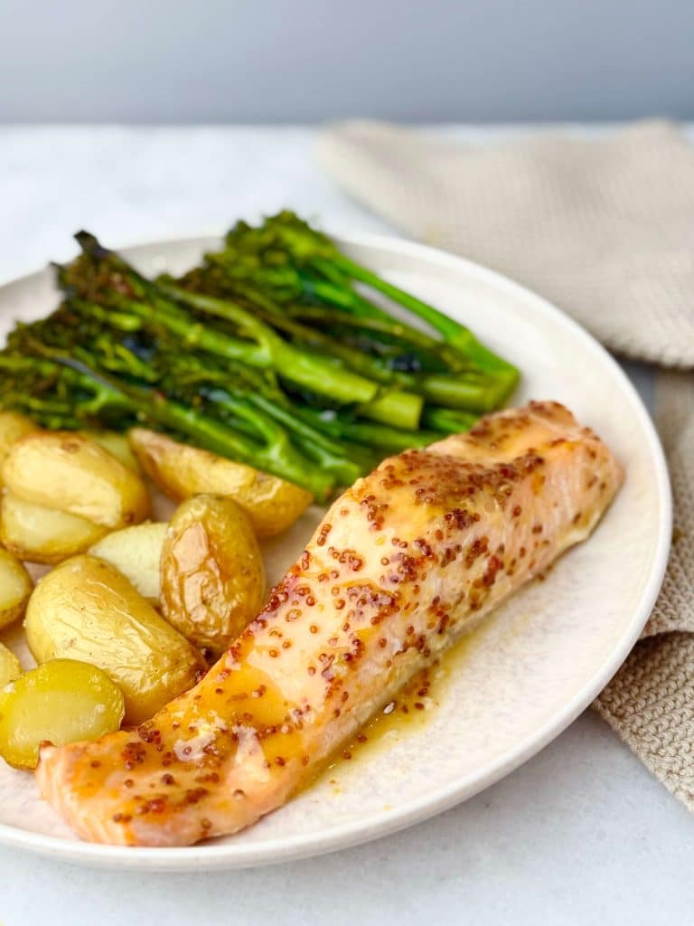 Close-up of a dinner plate with roasted baby potatoes, broccolini, and a honey mustard-glazed salmon fillet on a cream-colored ceramic plate.