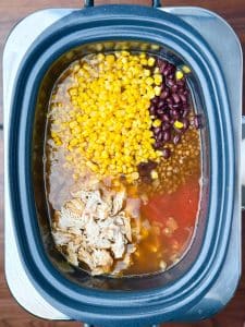 Slow cooker filled with shredded chicken, corn, black beans, lentils, and tomatoes just before simmering into tortilla soup.