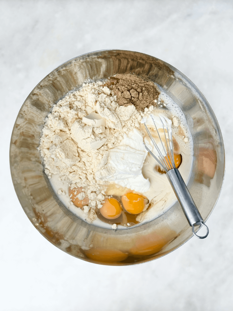 Mixing bowl with eggs, Greek yogurt, protein powder, and cardamom before whisking for baked oats.