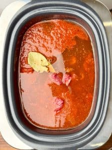 Beef chunks simmering in red chili broth with bay leaf in crockpot for birria soup.