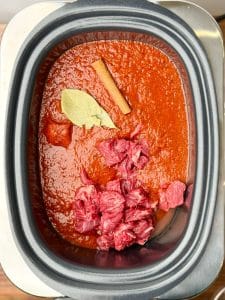 Raw beef chunks in crockpot with red chili sauce, cinnamon stick, and bay leaf for birria soup recipe.