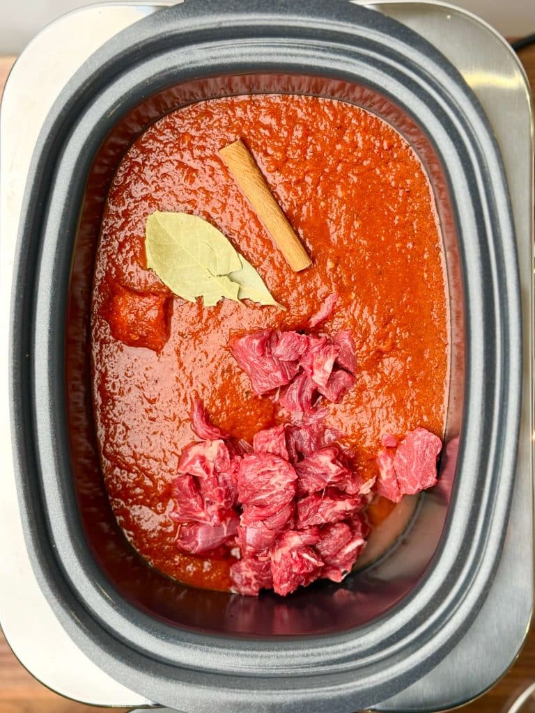 Raw beef chunks in crockpot with red chili sauce, cinnamon stick, and bay leaf for birria soup recipe.