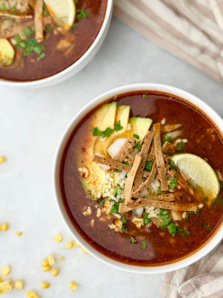 Close-up of birria soup with red chili broth, tortilla strips, avocado slices, and diced onion.