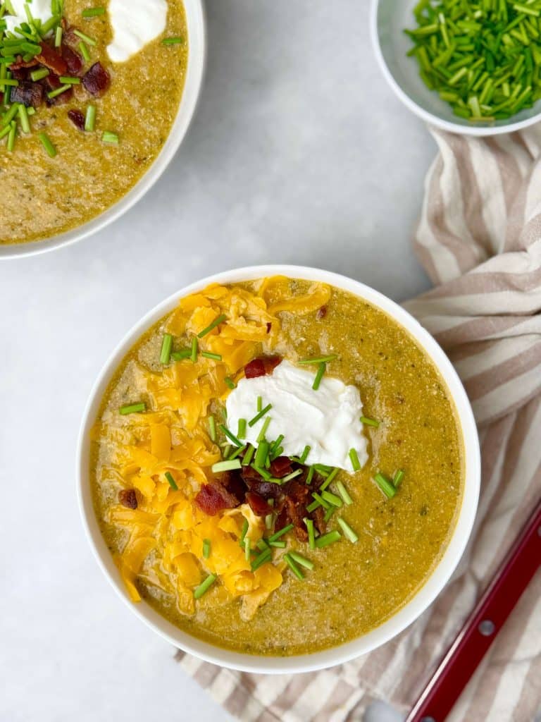 Two bowls of healthy crockpot broccoli cheddar soup with toppings on a white table