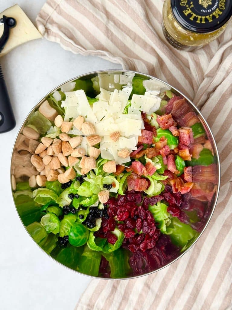 Mixing bowl filled with Brussels sprouts, bacon, almonds, dried cranberries, dried blueberries, and Manchego cheese shavings before tossing.