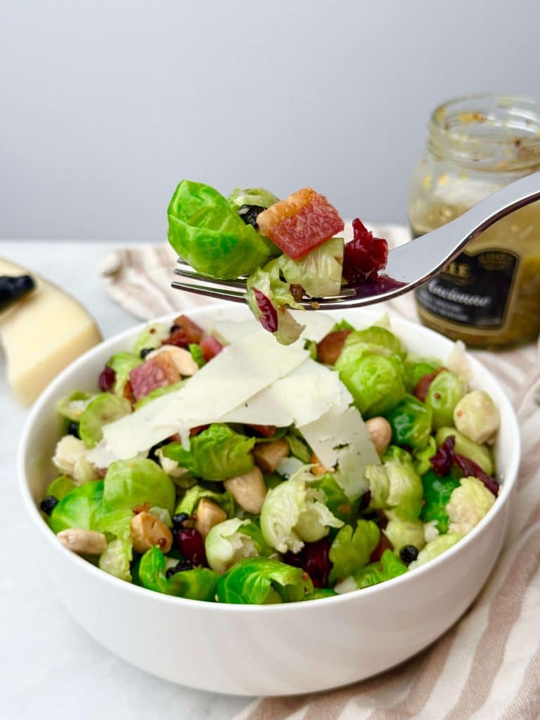 Fork holding a bite of brussel sprout salad with bacon, dried cranberries, blueberries, almonds, and Manchego cheese.