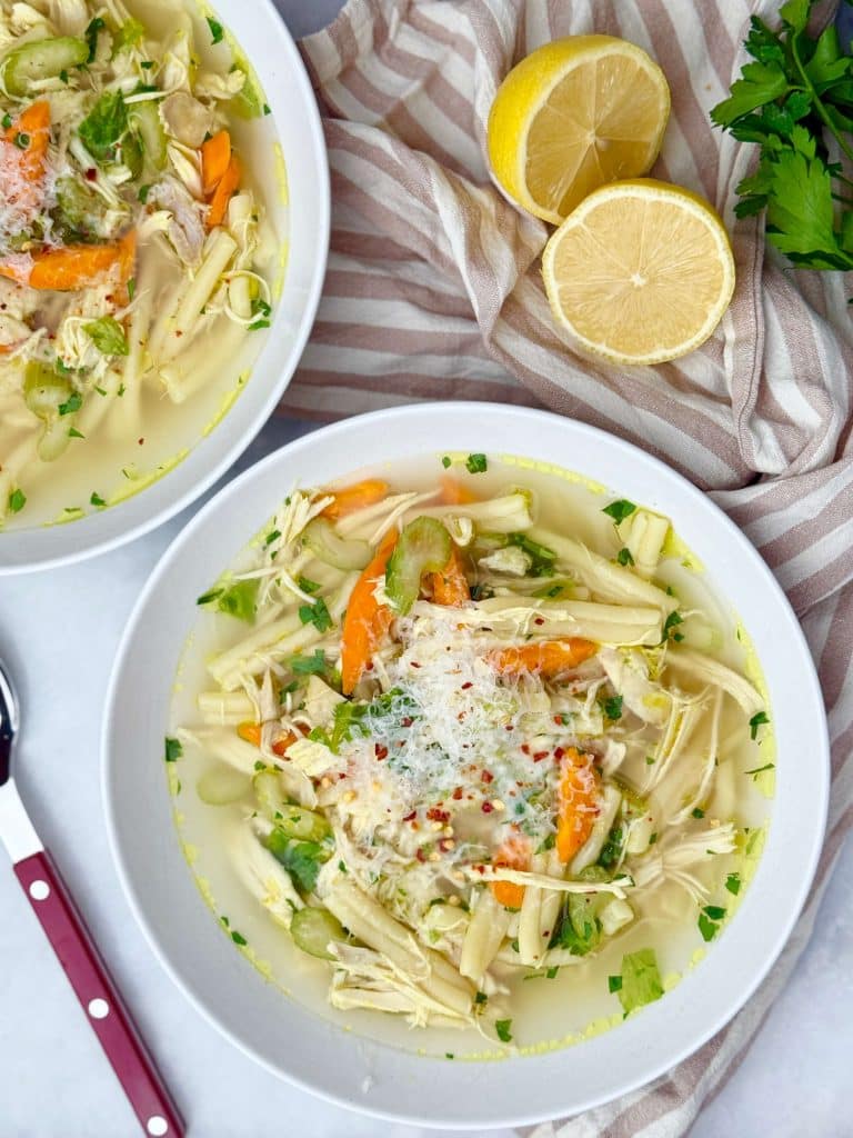 Two bowls of crockpot chicken noodle soup with lemon halves and fresh parsley on the side, topped with Parmesan and chili flakes.