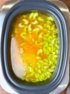 Golden chicken broth in a crockpot with sliced celery and carrots floating on top, ready to finish into chicken noodle soup.
