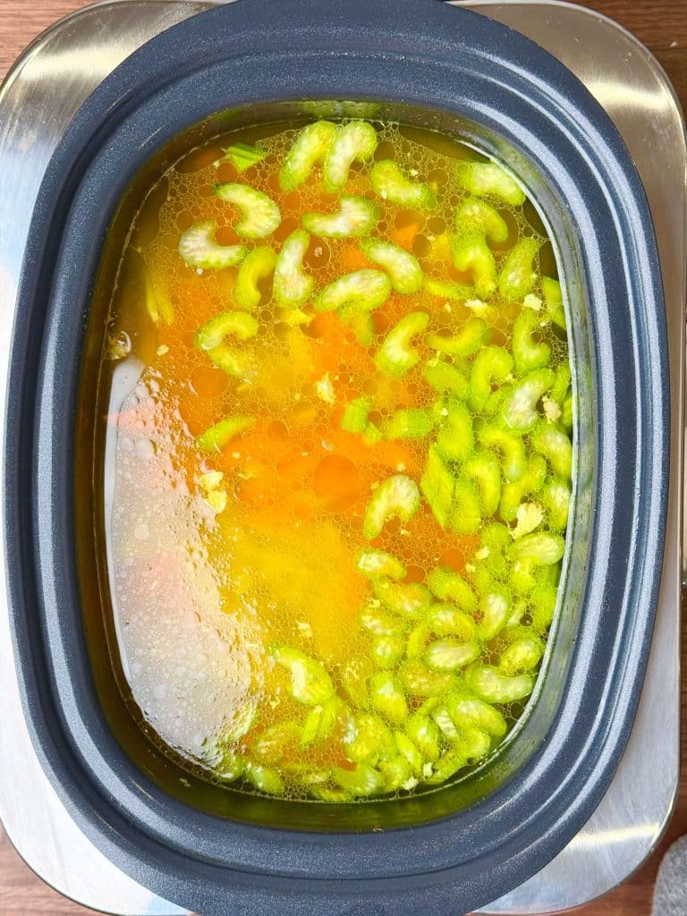 Golden chicken broth in a crockpot with sliced celery and carrots floating on top, ready to finish into chicken noodle soup.