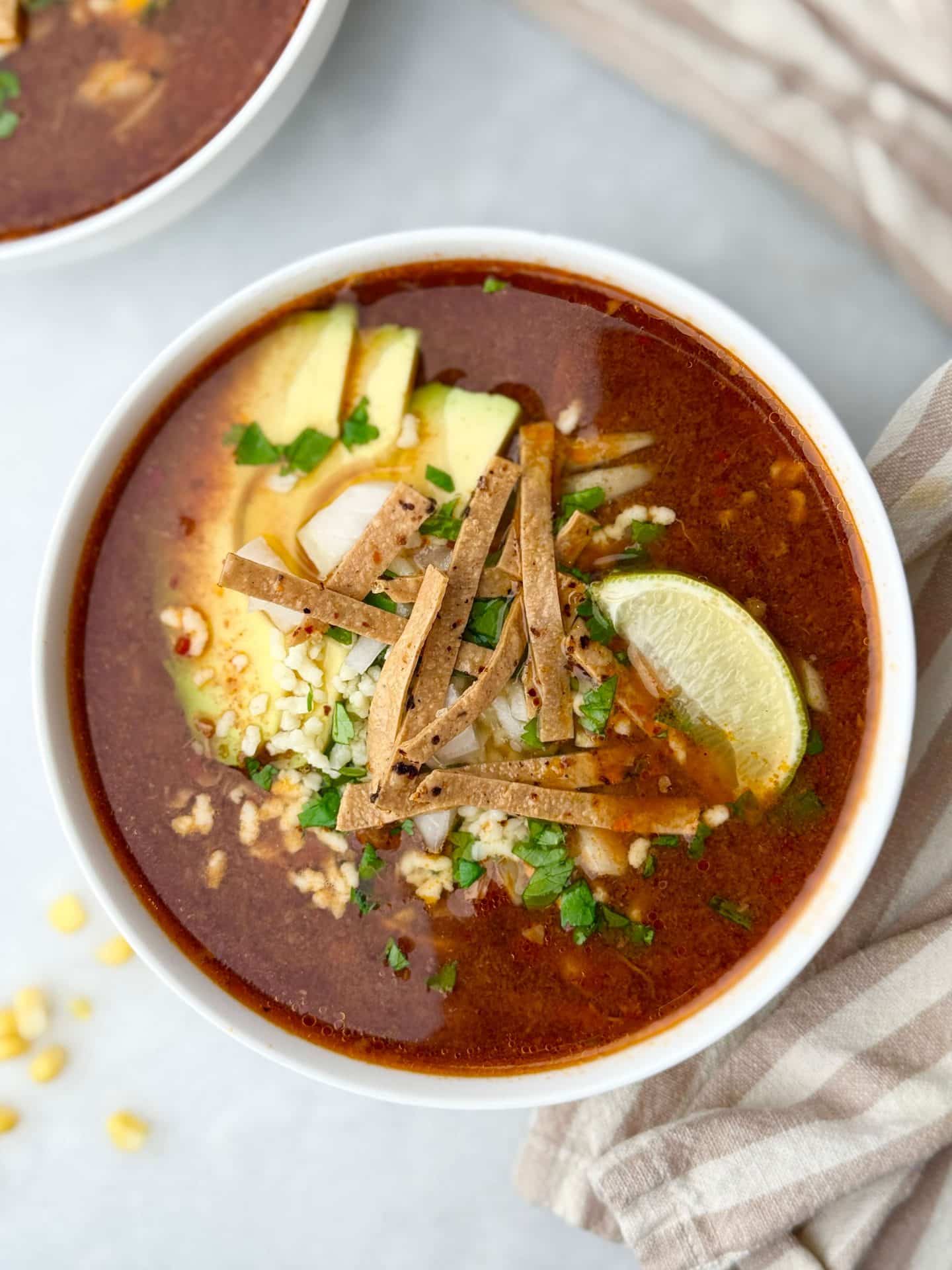 Overhead view of crockpot birria soup in a white bowl with tortilla strips, melted cheese, lime, and cilantro.