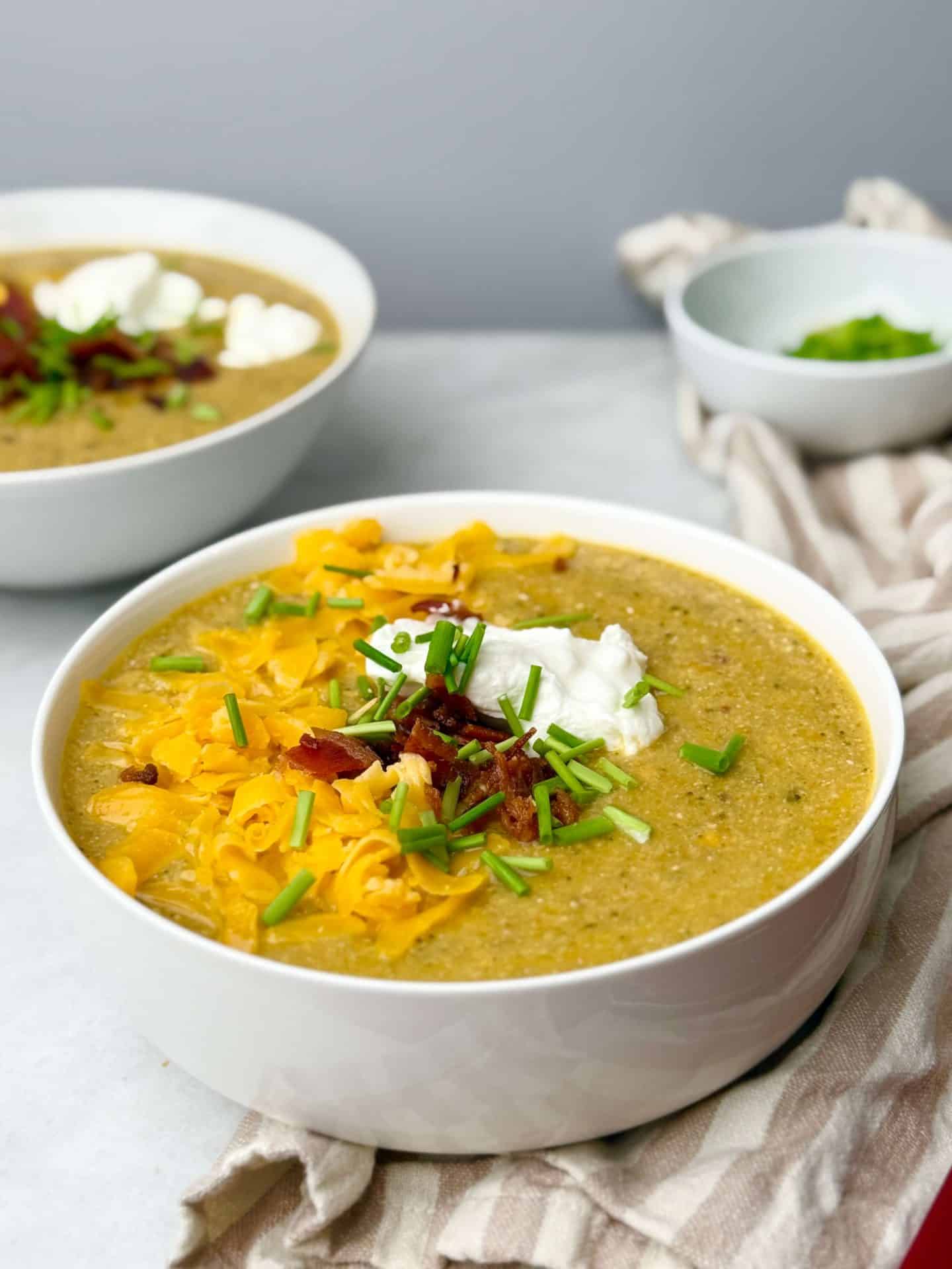 Bowl of high protein crockpot broccoli cheddar soup topped with cheddar, bacon, sour cream, and chives