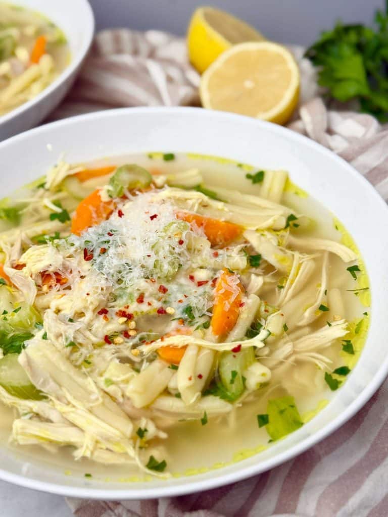 Bowl of crockpot chicken noodle soup topped with Parmesan cheese, fresh parsley, carrots, celery, and red pepper flakes, served with lemon on the side.