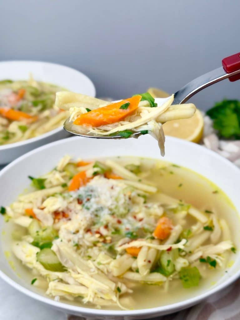 Close-up of a spoonful of chicken noodle soup with shredded chicken, carrot, and noodles, with a bowl of soup in the background.