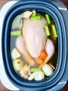 Whole chicken in a crockpot with onion, garlic, carrots, celery, ginger, and bay leaves, ready to cook into homemade chicken noodle soup.