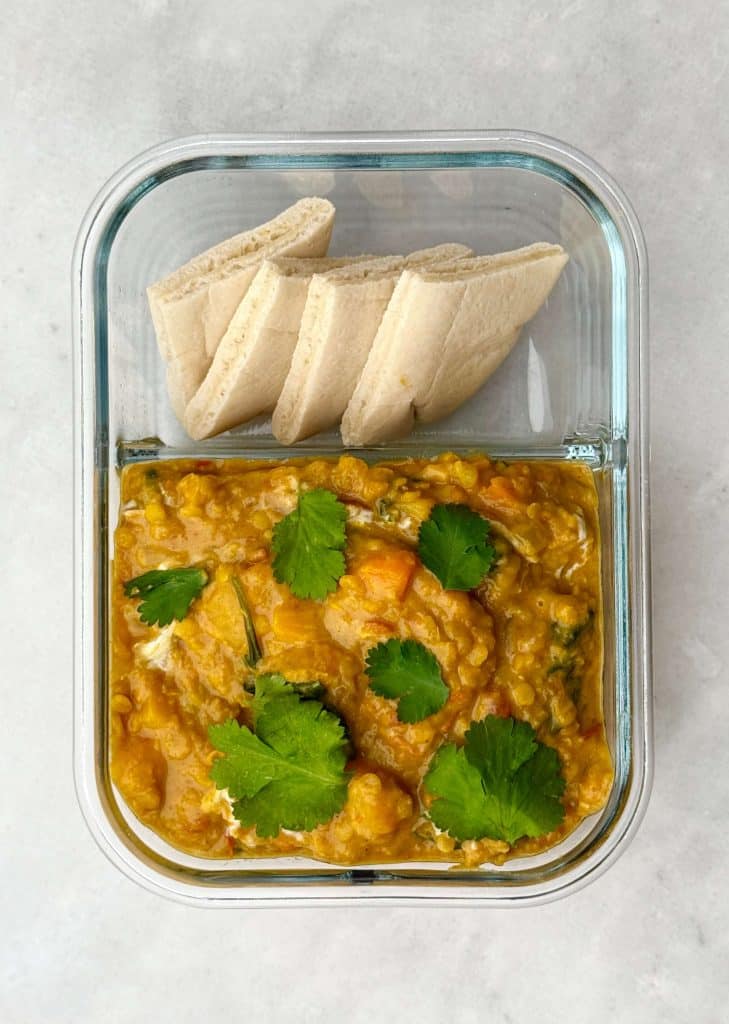 Meal prep container with crockpot butternut squash and red lentil curry, topped with cilantro and served with pita bread.