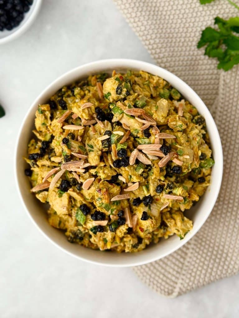 Overhead view of curry chicken salad topped with toasted almond slivers and dried blueberries in a white bowl.