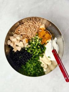 Ingredients for curry chicken salad in a bowl before mixing: chopped chicken, toasted almonds, curry powder, cilantro, jalapeño, blueberries, yogurt, and mayo.