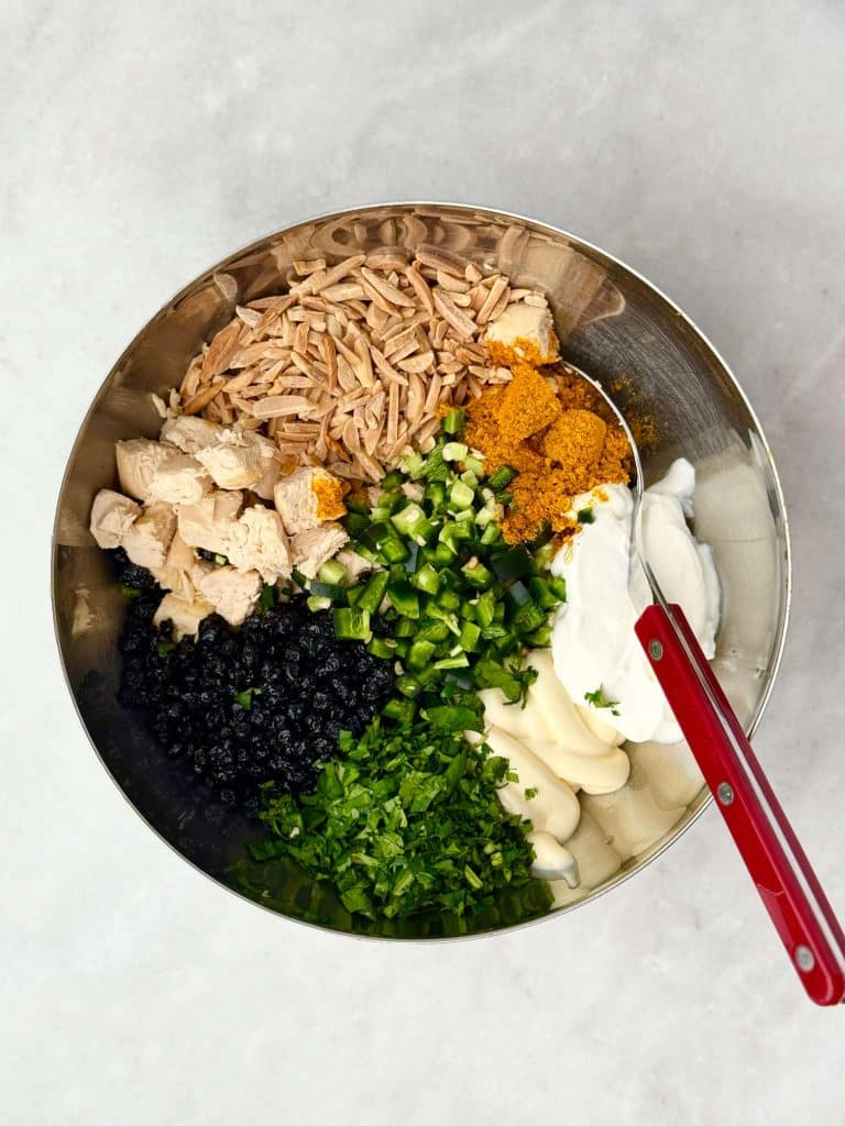 Ingredients for curry chicken salad in a bowl before mixing: chopped chicken, toasted almonds, curry powder, cilantro, jalapeño, blueberries, yogurt, and mayo.