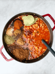 Spices including chili powder, smoked paprika, cumin, oregano, cocoa, and bay leaves on top of a simmering tomato base.