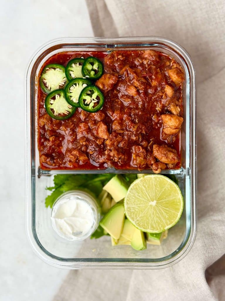 Chicken chili packed into a glass meal prep container with avocado, lime, cilantro, and a small cup of Greek yogurt.