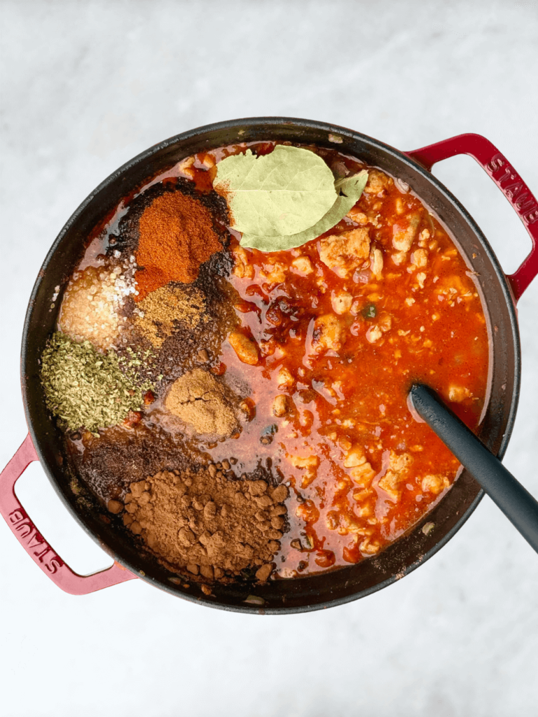 Spices including chili powder, smoked paprika, cumin, oregano, cocoa, and bay leaves on top of a simmering tomato base.