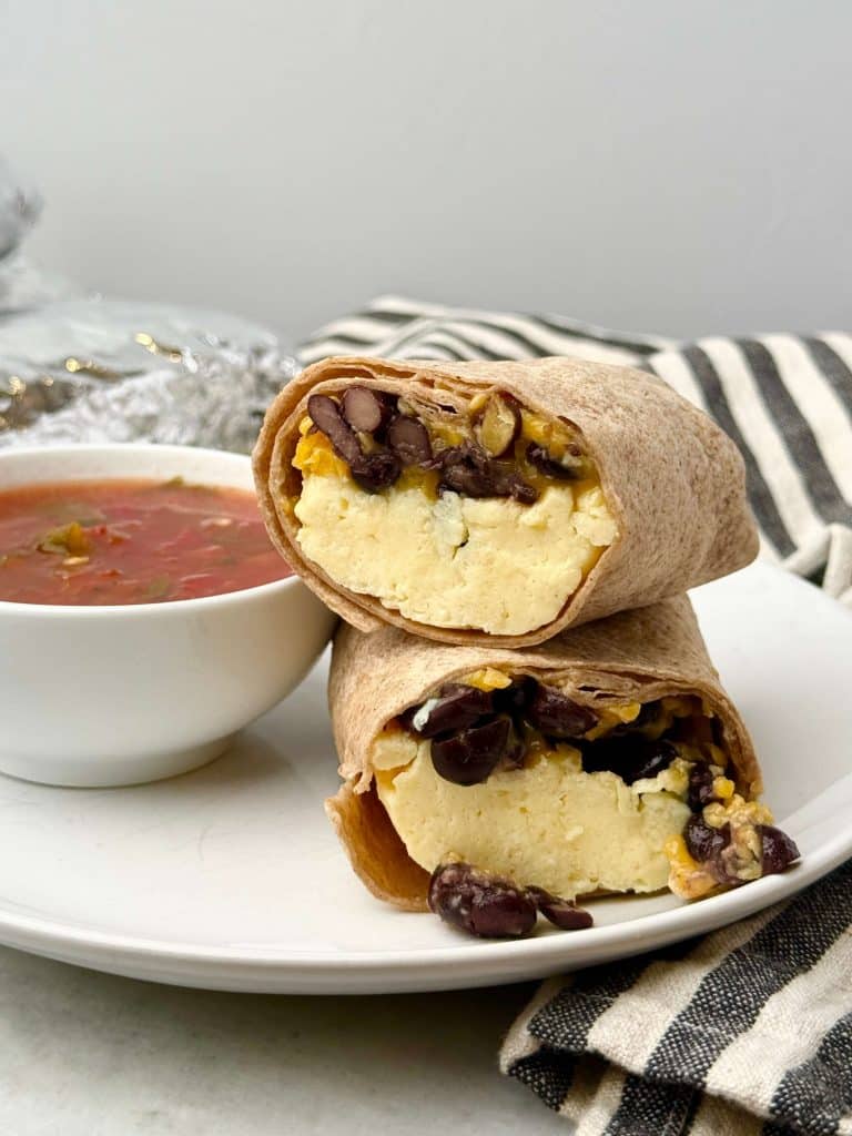 High protein freezer breakfast burrito cut in half on a white plate, filled with fluffy scrambled eggs, black beans, and melted cheddar, served with salsa.