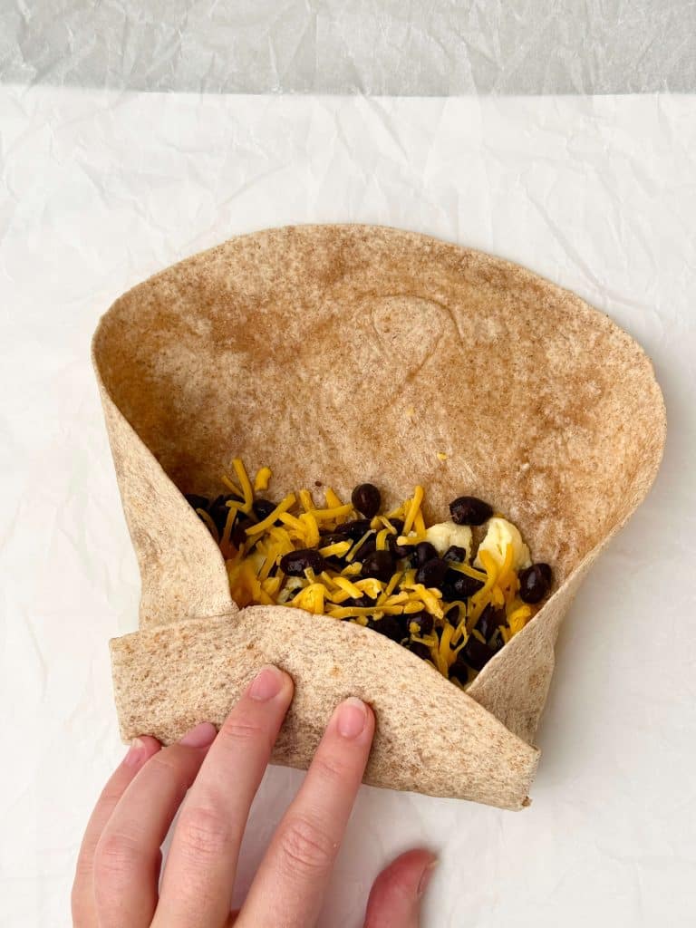 Hand folding a whole wheat tortilla filled with scrambled eggs, black beans, and shredded cheddar cheese to make a high protein freezer breakfast burrito.
