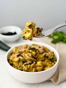 Fork lifting a bite of curry chicken salad with chicken, blueberries, and almonds over a white serving bowl.