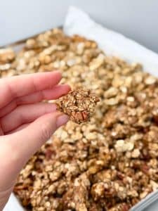 Hand holding a large crunchy cluster of homemade protein granola with pecans, with more granola cooling on a baking sheet in the background.