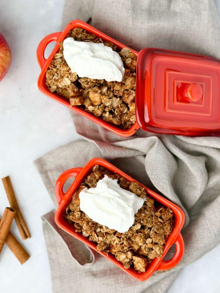 High protein apple crisp in red ramekins, topped with yogurt, styled with apples and cinnamon sticks for fall.