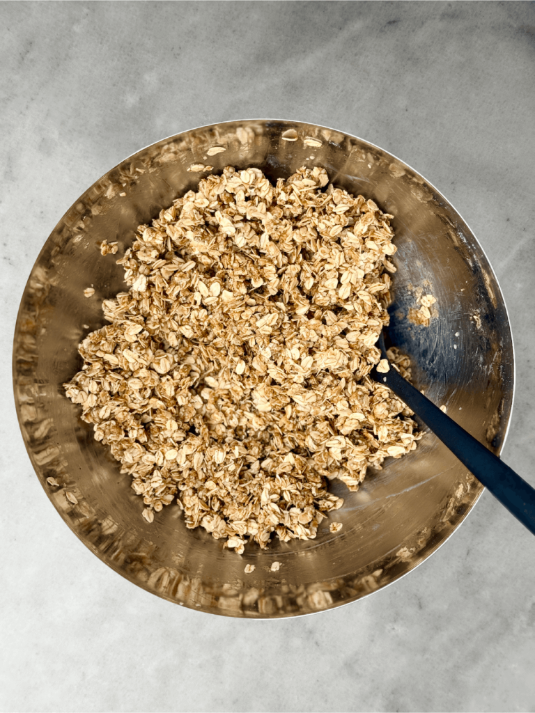 Rolled oats coated with almond butter mixture in a mixing bowl, ready to be baked into protein granola.