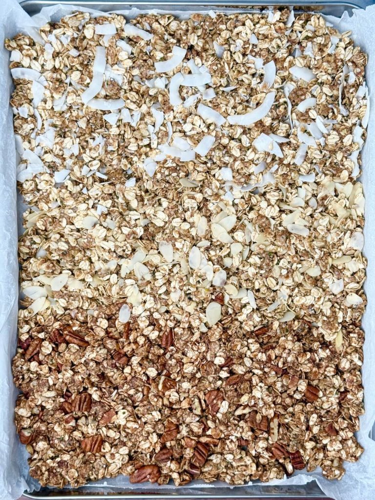 Sheet pan with three variations of homemade protein granola baked side by side — pumpkin spice with pecans, almond croissant with sliced almonds, and coconut cardamom with coconut flakes.