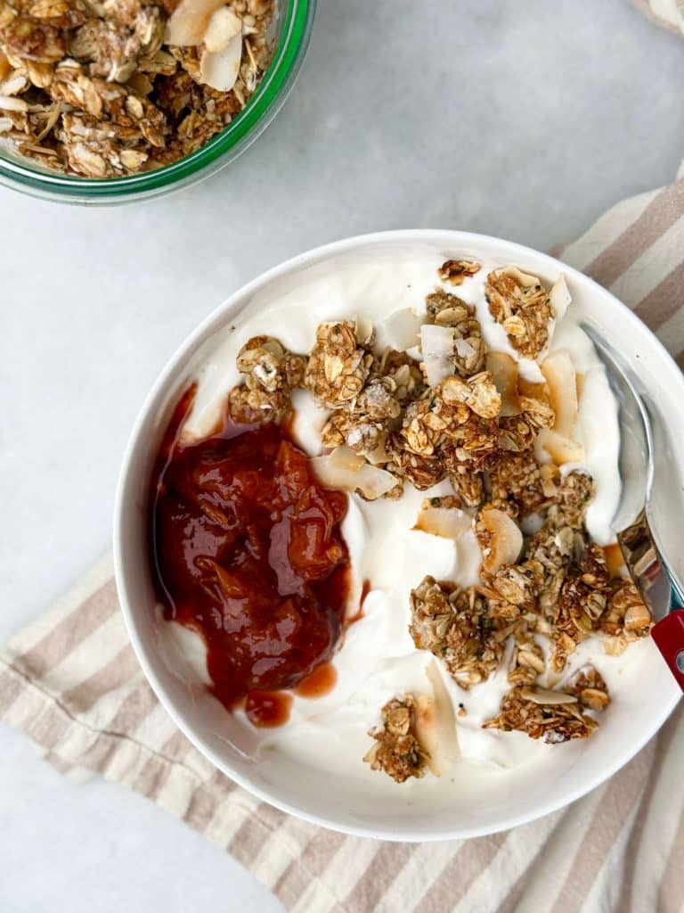 Bowl of Greek yogurt topped with homemade protein granola and strawberry jam, with extra granola in a jar nearby.