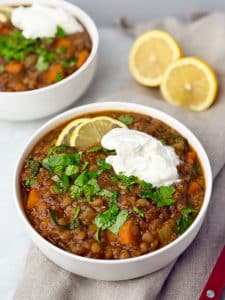 Close-up of hearty lentil soup in white bowls with lemon slices and a dollop of yogurt, garnished with chopped parsley.