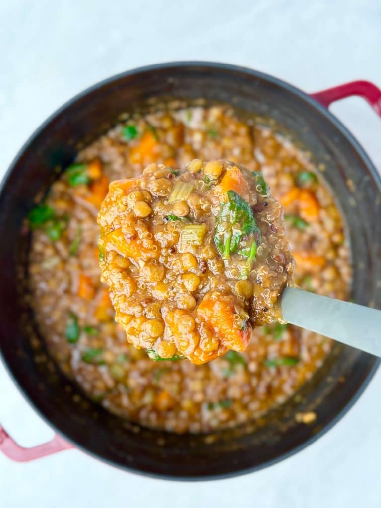 Spoonful of lentil soup with carrots, celery, and spinach lifted from a red Dutch oven filled with hearty vegetable soup.