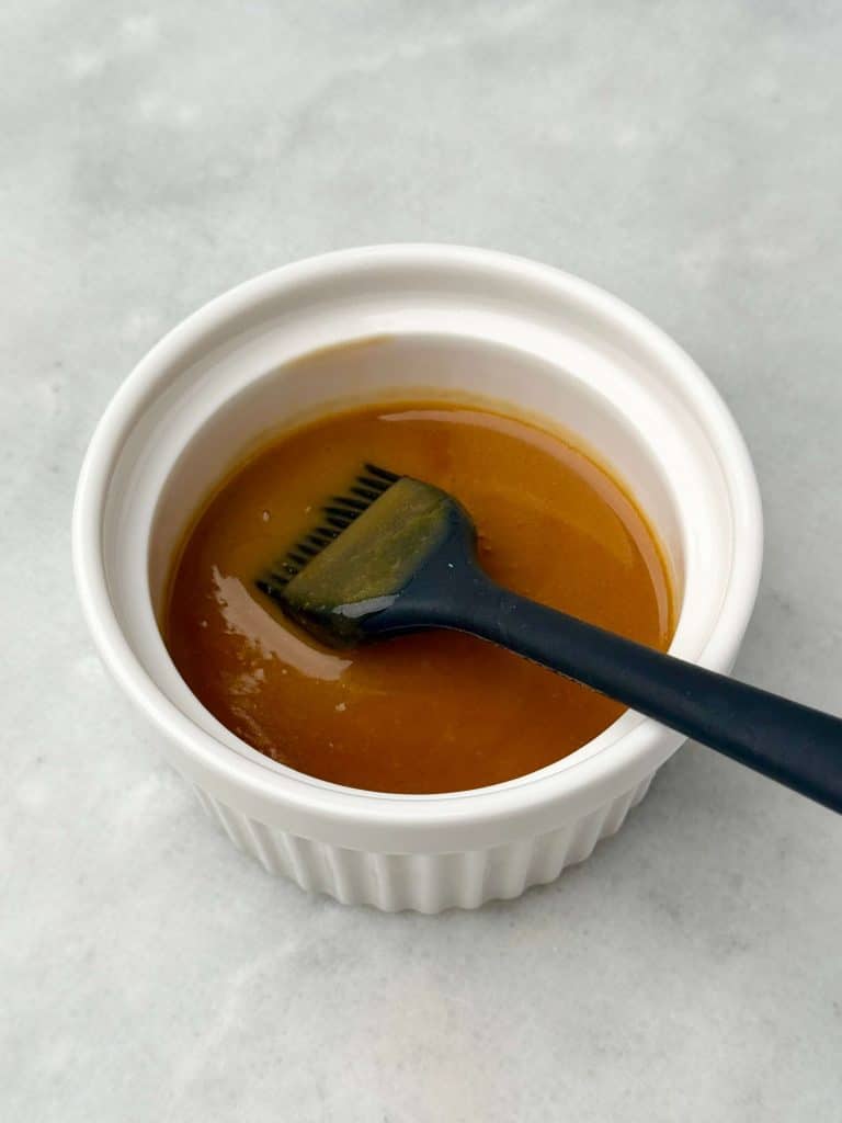 Glossy miso butter sauce in a small white ramekin with a black silicone brush